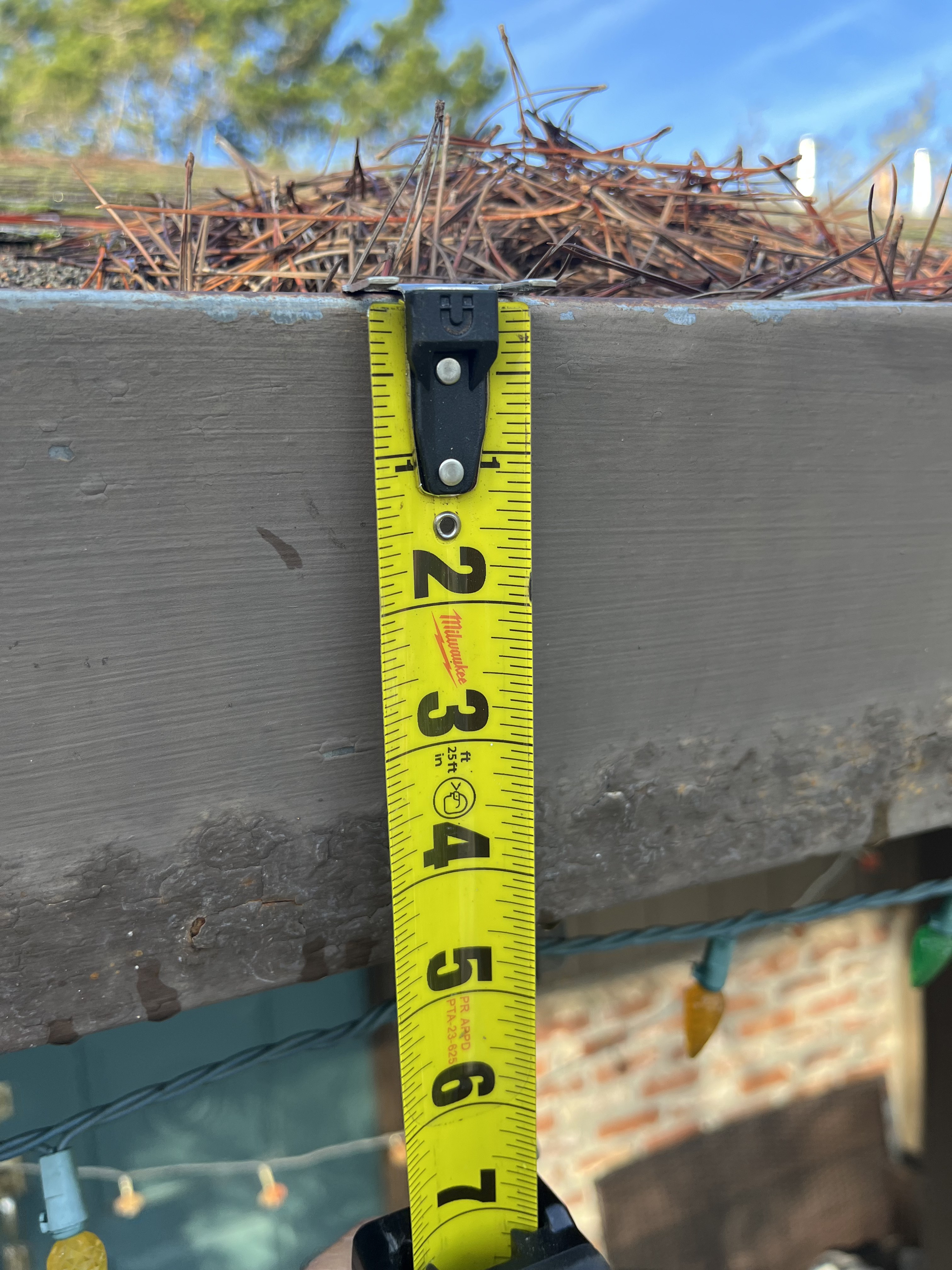 A Milwaukee tape measuring a five-inch fascia gutter packed with conifer debris — the kind of measurement that turns a subjective finding into a number you can share, dispute, or use.