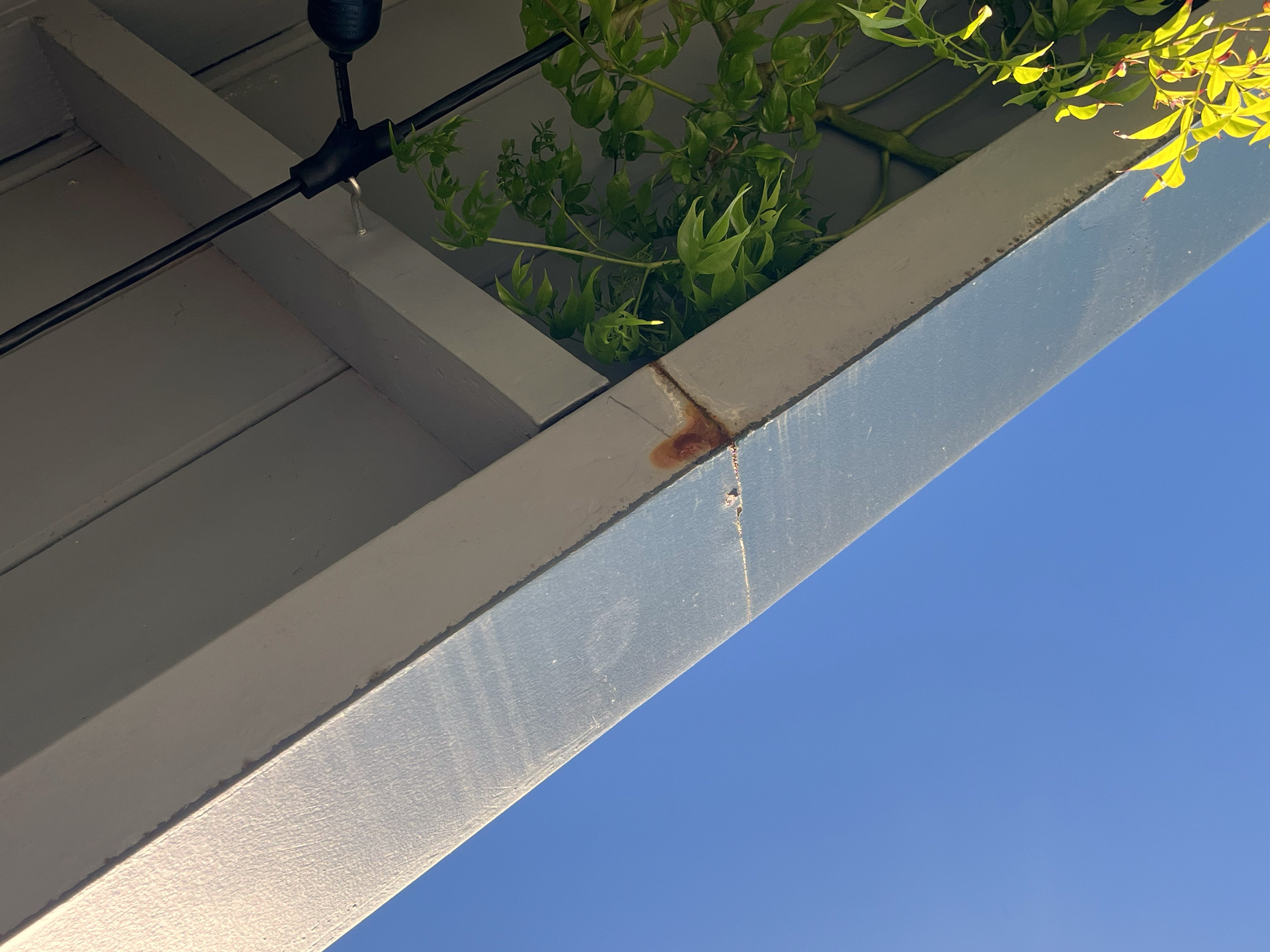 Rust-toned vertical streak originating from the bottom edge of a gutter where a rear seam has failed — water running down the back of the channel and wrapping around the underside