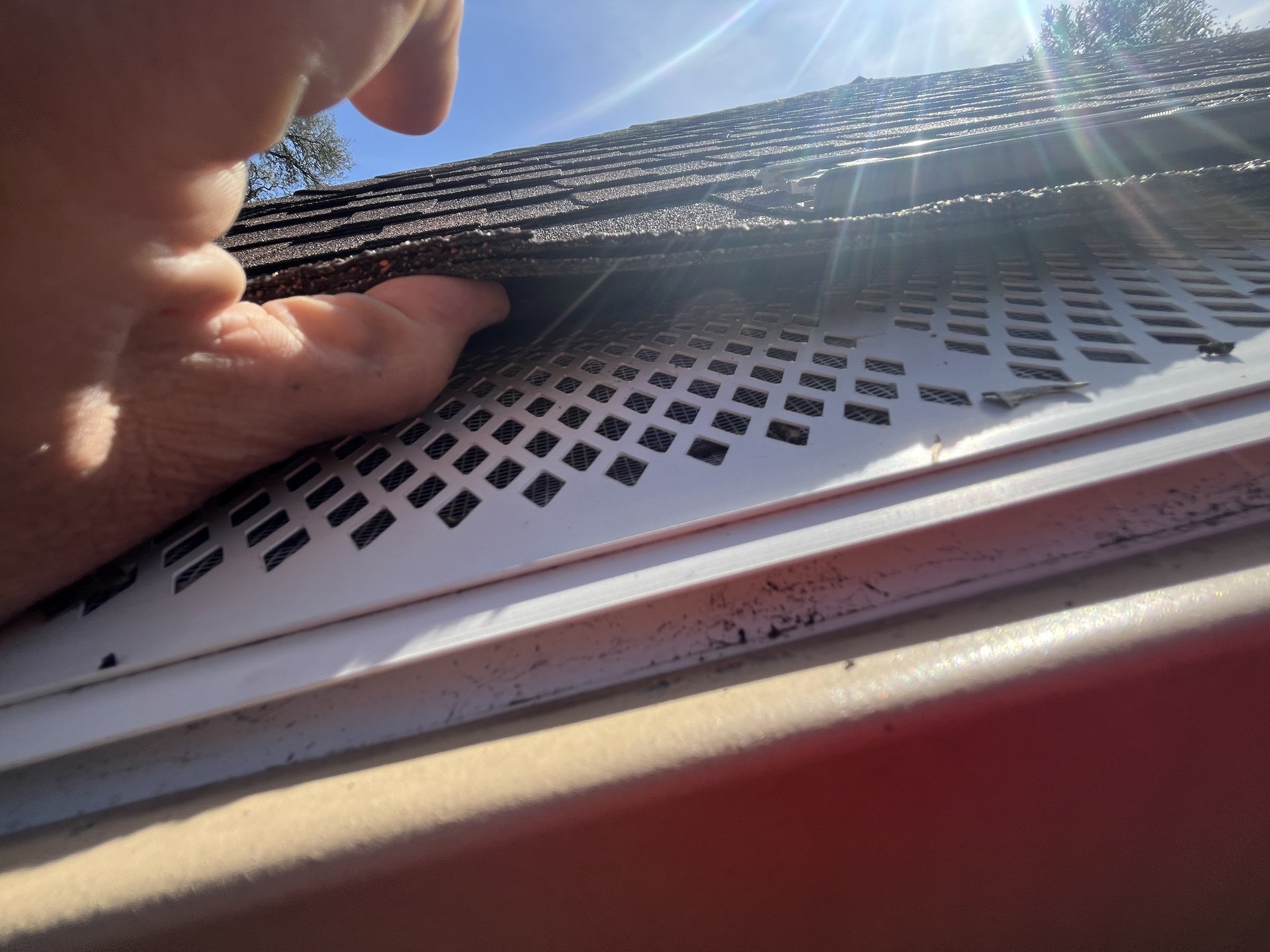 A hand lifting a perforated metal gutter cover — the reveal under your cover is what would otherwise stay hidden above your eye line indefinitely.