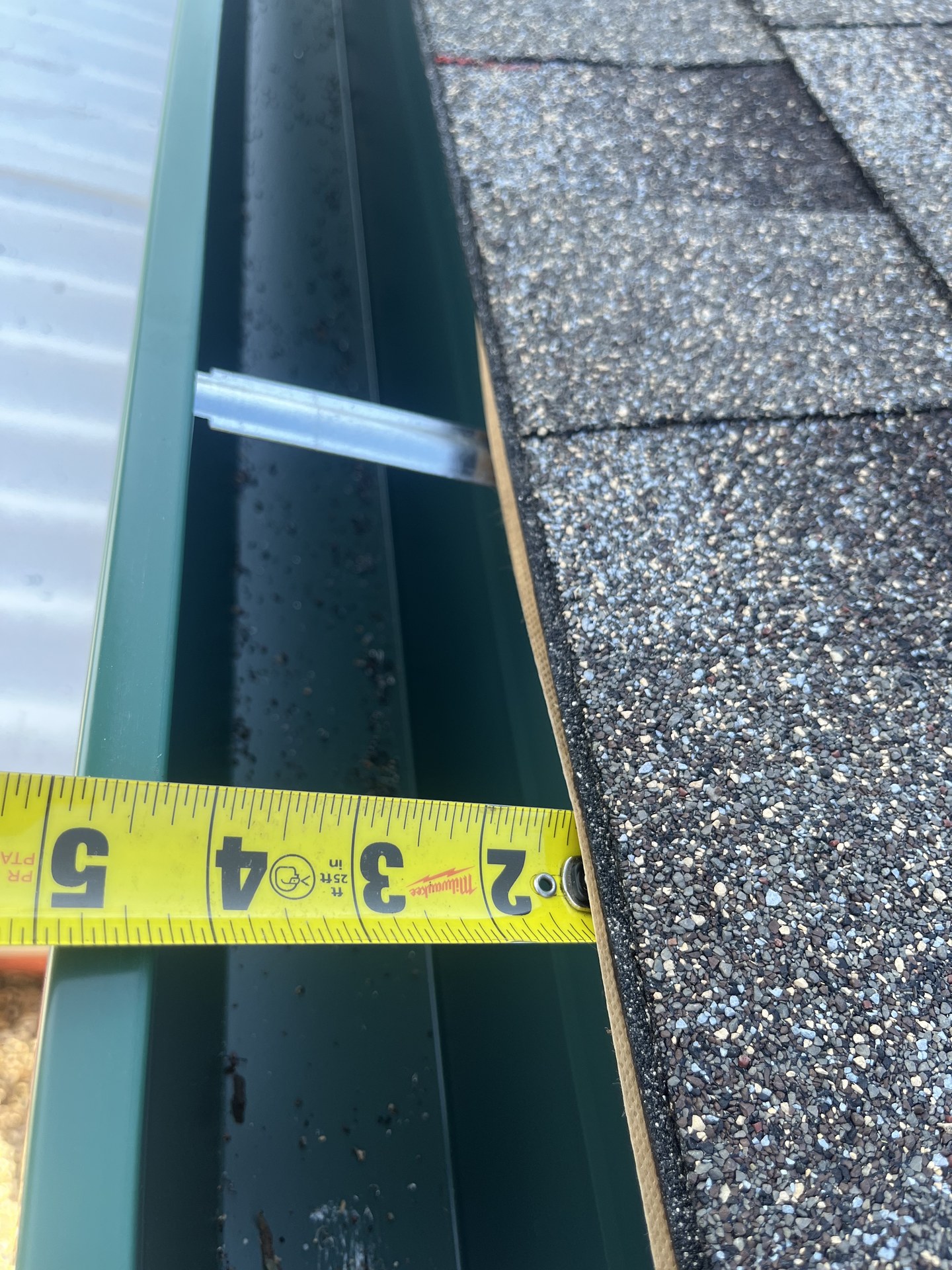 Milwaukee tape measuring four-inch narrow-profile gutter width at the galvanized T-bar bracket — the contact hardware where dissimilar metals meet inside the channel. Northern California field inspection.